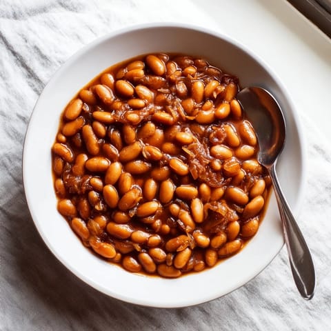 A spoon lifts tender baked beans from a Dutch oven, revealing molasses-rich sauce beside grilled sausages and cornbread.